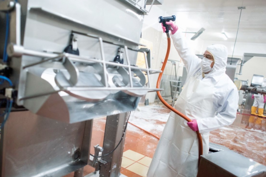 Ein Arbeiter in Schutzkleidung – weißer Anzug, Maske und rosa Handschuhe – reinigt Industrieanlagen in einer Lebensmittelproduktion mit einem Schlauch während sich auf dem Boden etwas Schaum bildet.
